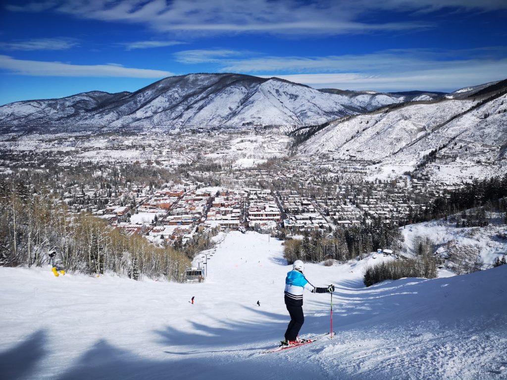 A skier at the top of a slope views a snowy ski run and the town of Aspen below