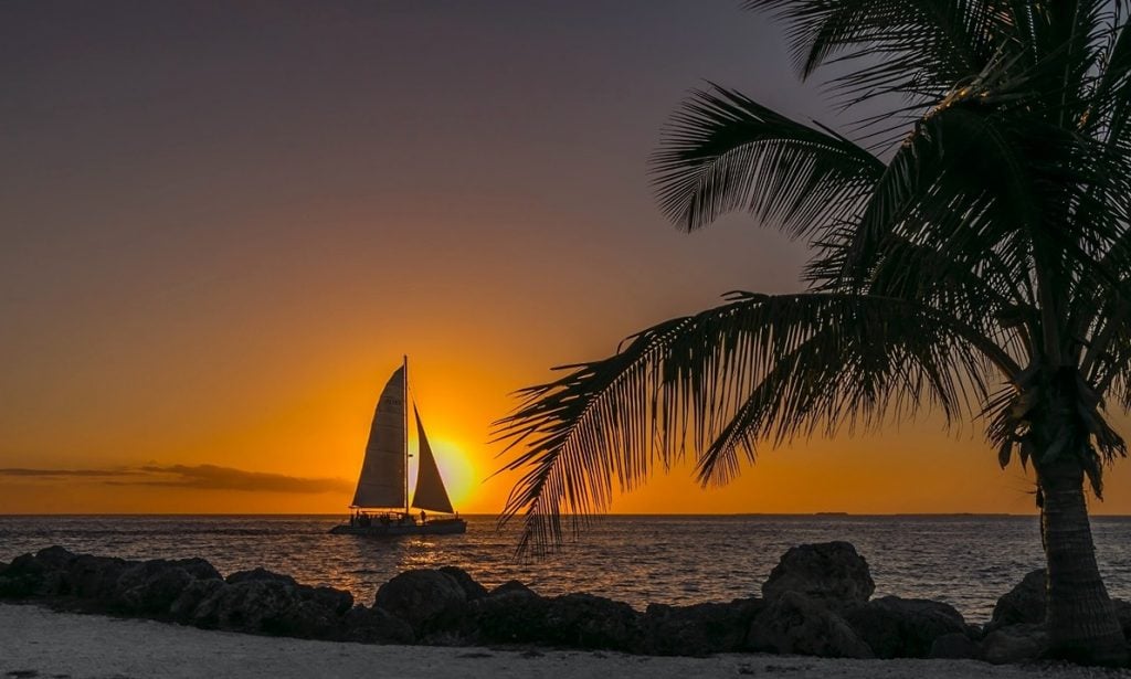 A sailboat and palm tree in the foreground, as the sun sets behind the ocean horizon in the Florida Keys