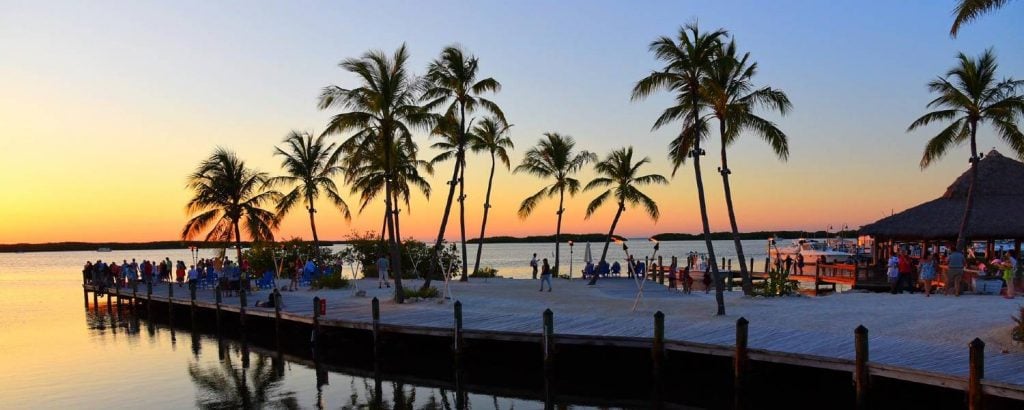 A group of people look at at the sunset from a sandy pier next to palm trees and tiki torches in the Florida Keys