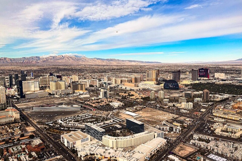 An aerial view of the Las Vegas Strip in daylight, with the Sphere visible and mountains under dramatic clouds behind