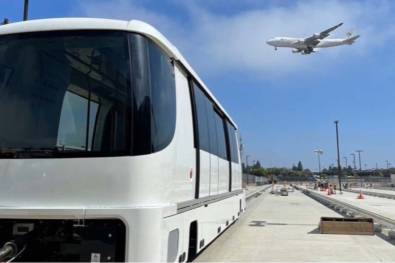 A white LAX Automated People Mover train parked on the guideway at the maintenance facility, with an airliner on final approach in the sky behind it