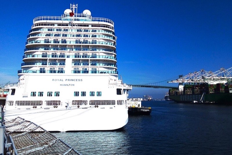A cruise ship berthed at the World Cruise Center in San Pedro under a deep blue sky, with the Vincent Thomas Bridge and port cranes behind it