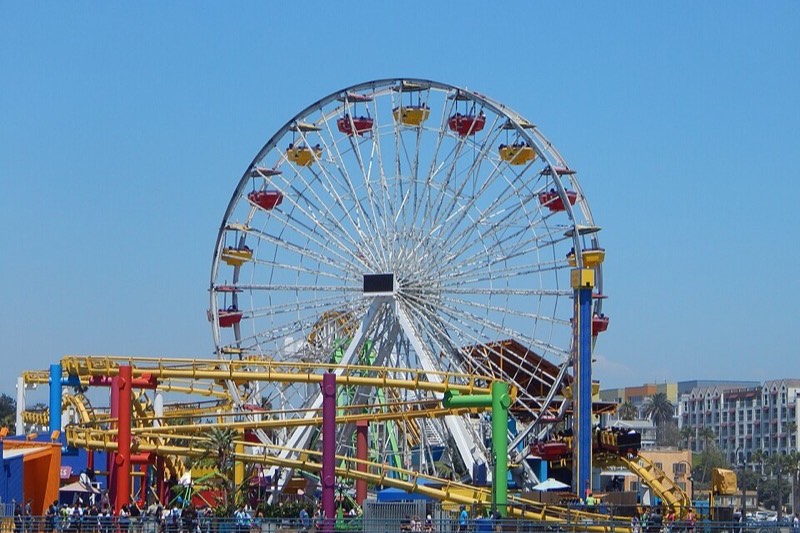 The Pacific Wheel ferris wheel and roller coaster on Santa Monica Pier against a deep blue sky, with crowds on the pier deck